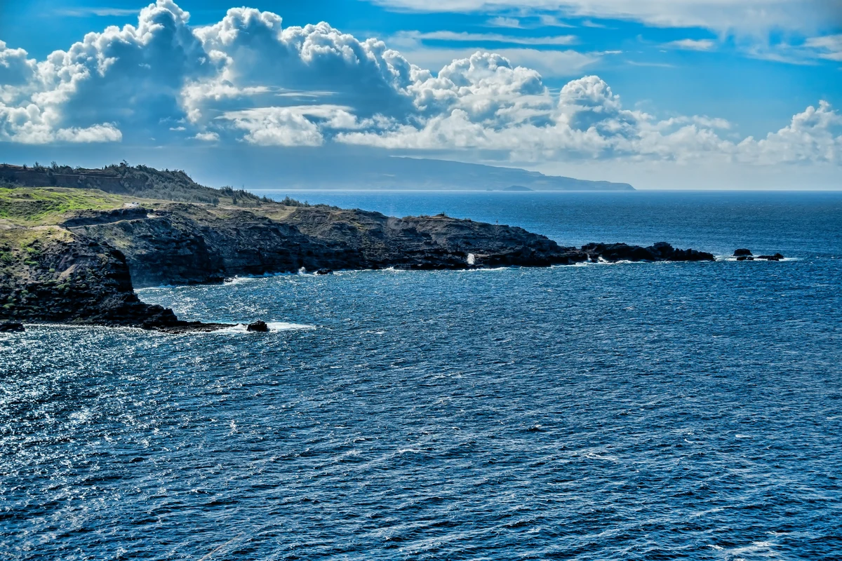 Costa rochosa de Maui junto ao mar azul, sob nuvens dispersas.